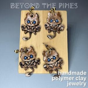 May include: Four pairs of handmade polymer clay earrings featuring a brown and white creature with blue eyes and wings. The earrings are displayed on a wooden board with the text "BEYOND THE PINES" and "handmade polymer clay jewelry".