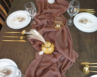 May include: A brown table runner with a textured surface is laid out on a dark wood table. The runner is decorated with dried pampas grass in a white vase and gold candle holders. There are white plates, gold silverware, and wine glasses set on the table.
