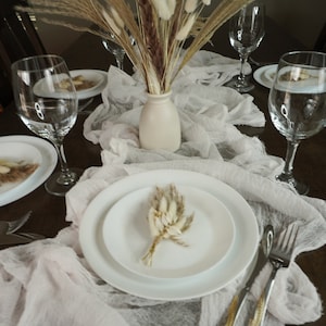May include: A white table runner with a white vase filled with dried pampas grass. Two white plates with dried pampas grass on top are set on the table. Two wine glasses and a fork and knife are also on the table.
