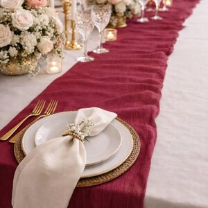 May include: Elegant table setting with a burgundy runner, white plates, gold silverware, and floral centerpieces. The table is set for a special occasion, with candles and wine glasses adding to the ambiance.