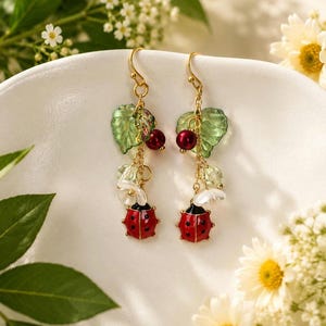 May include: Gold-tone drop earrings with ladybug charms. The earrings feature red and black ladybug pendants, green leaf accents, and red bead details. White flower accents and gold hooks complete the design.