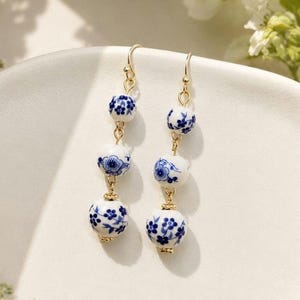 Op de afbeelding: Een paar oorbellen met drie in grootte variërende witte keramische kralen met blauwe bloemmotieven. De kralen zijn verbonden door goudkleurige metalen schakels en hangen aan goudkleurige oorhaken. Elke oorbel toont een consistent ontwerp van kleine blauwe bloemen en fijne ranken die op de bolvormige oppervlakken zijn geschilderd.