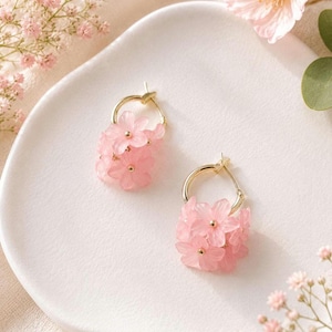 May include: Gold hoop earrings, each featuring a cluster of pink flowers. The earrings have a gold-coloured hoop design, with a collection of small, translucent pink flowers. The earrings are displayed on a white surface, with a soft, neutral background.