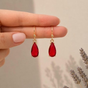 Puede incluir: Un par de pendientes colgantes de tono dorado con piedras rojas facetadas en forma de lágrima. Cada pendiente cuelga de un fino gancho de alambre dorado, mostrando un tono carmesí claro y vibrante con un acabado pulido y reflectante.