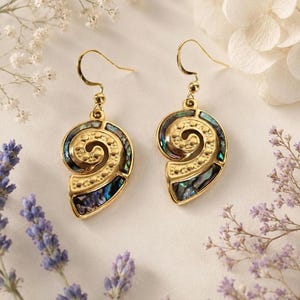 May include: A pair of gold-toned drop earrings featuring a spiral shell-inspired design. Each earring is inlaid with iridescent abalone shell fragments and accented with a textured dotted pattern along the centre curve, suspended from gold hooks.