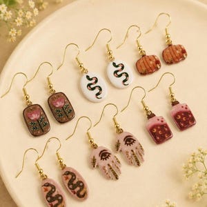 May include: A collection of five pairs of dangle earrings with gold-toned hooks. Designs include white oval snake charms, brown rectangular floral motifs, pink hand-shaped charms with eye symbols, red potion bottles with moon icons, and orange pumpkin shapes.