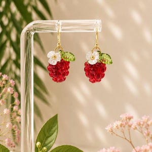 May include: A pair of gold hoop earrings featuring red beaded raspberry charms. Each charm is adorned with a white flower and a green leaf. The earrings are displayed on a clear stand against a soft background.