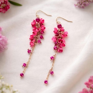 May include: Dangle earrings with a floral design. The earrings feature clusters of small, bright pink flowers and small pink beads hanging from gold-coloured hooks and chains. The earrings are set against a white background.