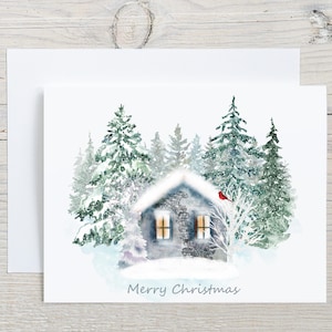 May include: A white greeting card featuring a watercolor illustration of a stone cottage covered in snow, surrounded by evergreen trees. A small red cardinal sits on a bare tree branch. The text Merry Christmas is printed in gray at the bottom.