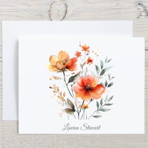 May include: A white rectangular greeting card featuring a watercolor illustration of orange and yellow flowers with green foliage. The name Lauren Stewart is printed in a dark gray serif font at the bottom center of the card, which sits on an envelope.
