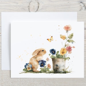 May include: Watercolor illustration of a bunny with blue, yellow, and pink flowers in a metal pot. A yellow butterfly flutters nearby. The artwork is on a white card with an envelope, perfect for Easter or spring.