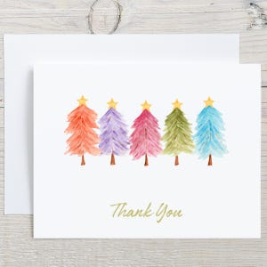May include: A white greeting card featuring five watercolor-style Christmas trees in orange, purple, pink, green, and blue, each topped with a yellow star. The text Thank You is printed in gold cursive script below the trees, with a matching envelope.
