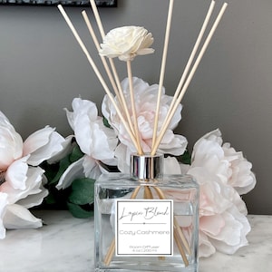 May include: A clear glass bottle with a silver lid and wooden sticks, filled with a clear liquid. The bottle has a label that reads "Lapin Blond Cozy Cashmere Room Diffuser 4 oz / 120 ml". The bottle is surrounded by white and pink flowers.