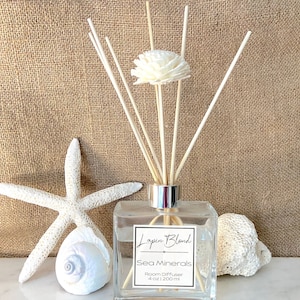 May include: A clear glass bottle with a silver lid and wooden sticks, filled with a clear liquid. The bottle has a label that reads "Layla Blond Sea Minerals Room Diffuser 4 oz | 200 ml". The bottle is surrounded by seashells and a starfish on a beige hessian background.