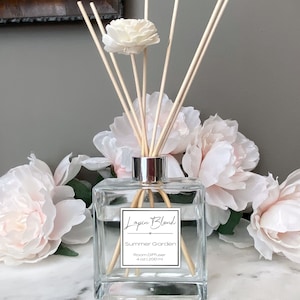 Op de afbeelding: Een transparante glazen diffuser met een zilveren dop en houten stokjes. De fles is gevuld met een heldere vloeistof en heeft een etiket met de tekst "Lapin Blend Summer Garden Room Diffuser 4 oz / 120 ml". De fles is omringd door witte bloemen.