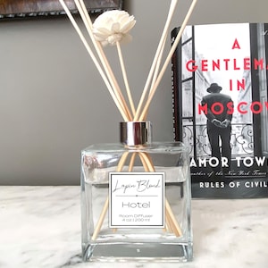 May include: A clear glass bottle with a silver lid and wooden sticks, filled with a clear liquid. The bottle has a label that reads "Lapin Blond Hotel Room Diffuser 4 oz | 120ml". The bottle is sitting on a white marble surface next to a book titled "A Gentleman in Moscow" by Amor Towles.
