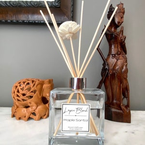 Puede incluir: Un difusor de ambiente de vidrio transparente con palitos de madera y una flor blanca. La botella tiene una etiqueta que dice "Logan Blend Maple Santal Room Diffuser 4 oz / 120 ml".