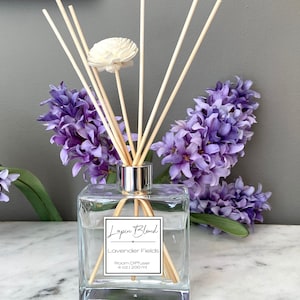 Puede incluir: Un difusor de ambiente de vidrio transparente con varillas de madera y un acento floral blanco. La etiqueta dice "Lapin Blanc Lavender Fields 4 oz / 200 ml". Flores de jacinto moradas están dispuestas alrededor del difusor.