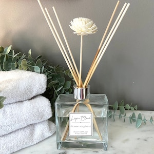 以下が含まれることがあります： 木製のリードと白い花が付いた、透明な正方形のガラスディフューザーボトル。「Lapin Blend Rosemary Mint」と書かれたラベル。ボトルは、白いタオルと緑の背景がある大理石の表面に置かれています。ディフューザーには120mlが含まれています。