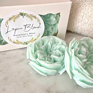 Pode incluir: Dois sabonetes de leite de cabra verde claro em forma de flor numa caixa branca com um rótulo que diz "Lapin Blond' Rosemary Mint Goat Milk Soap".