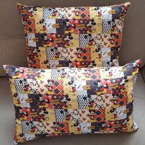 May include: Two decorative throw pillows with a patchwork design featuring Mickey Mouse. The pillows are yellow, brown, black, red, and white with a repeating pattern of Mickey Mouse in various poses. The text "Mickey Mouse" is repeated throughout the design.