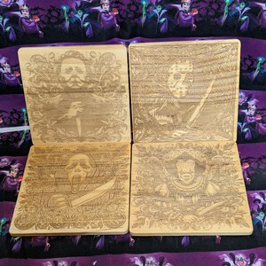 May include: Four square wooden coasters with engraved portraits of horror movie characters. Each coaster features a different character, including a man with a knife, Jason Voorhees, Ghostface, and the Evil Queen, surrounded by decorative flourishes.