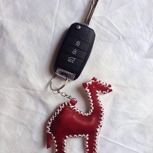 Op de afbeelding: Een rode leren kameel sleutelhanger met witte stiksels. De sleutelhanger is bevestigd aan een zwarte autosleutel met een zilveren sleutelmes. De sleutel heeft vier knoppen: ontgrendelen, vergrendelen, kofferbak en vasthouden.