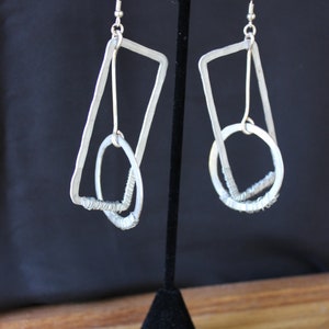 Puede incluir: Un par de pendientes de plata con un diseño geométrico. Cada pendiente tiene un rectángulo grande con un círculo más pequeño en su interior. Los círculos están hechos de alambre en espiral.