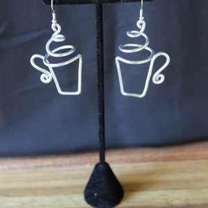 Puede incluir: Un par de pendientes de alambre plateado con forma de taza de café con un diseño de espiral en la parte superior.