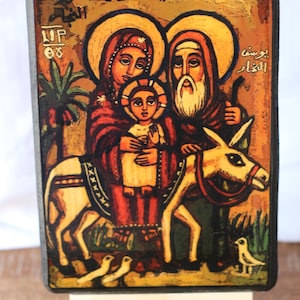 May include: A colorful painting on wood depicting the Holy Family, Mary, Joseph, and baby Jesus, riding a donkey. The painting is framed in black and mounted on a small wooden easel. The text in the image reads "The Flight into Egypt".