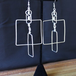 May include: Silver-toned wire earrings with a geometric design. Each earring features a square and a rectangular shape, connected by wire loops and a hook. The earrings are displayed on a black stand against a dark background.