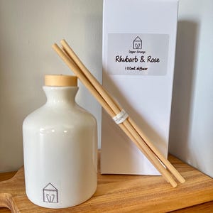 May include: A white ceramic diffuser bottle with a wooden cap, wooden reeds, and a white box labelled "Rhubarb & Rose 100ml diffuser." The items are displayed on a wooden board, creating a spa-like aesthetic.