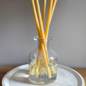 May include: A clear glass diffuser bottle with yellow reeds, filled with a clear liquid. The bottle sits on a white, round tray. The reeds are arranged in a fan shape, and the bottle has a small label with a house graphic.