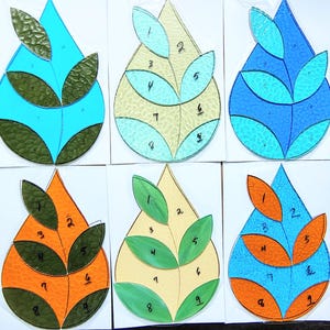 May include: Six colorful stained glass patterns in a teardrop shape, each with leaf-like designs and numbered sections. The color schemes include blue, green, orange, and yellow, with dark green and orange accents. The designs are on white cards.