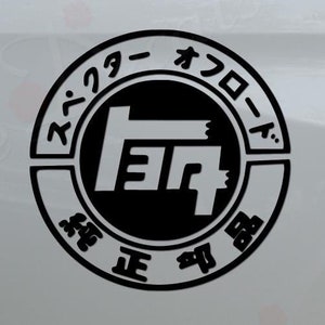 May include: Black and white decal of the Toyota logo with Japanese text around the outside. The decal is on the back of a white pickup truck.
