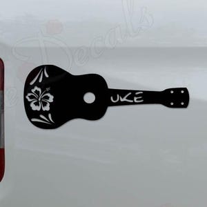 May include: Black ukulele-shaped decal with a floral design and the word "UKE" in white. The decal is affixed to the side of a white vehicle. The ukulele design features a hibiscus flower.