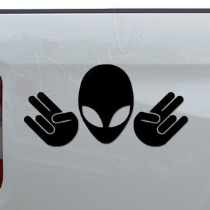 May include: Black and white alien sticker with two hands making the 'peace' sign on a white truck tailgate.
