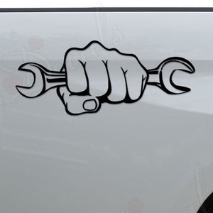 May include: A black and white decal of a fist holding a wrench. The decal is applied to the back of a white pickup truck.
