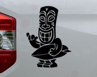 Puede incluir: Calcomanía en blanco y negro de una figura tiki sosteniendo una tabla de surf. La calcomanía está en la parte trasera de una camioneta blanca.