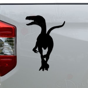 May include: A black dinosaur decal, in a running pose, is affixed to the side of a white truck. The decal is a silhouette of a raptor with its mouth open. The truck's taillight and bumper are visible.