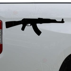 May include: A black decal of an AK-47 assault rifle is affixed to the white side of a vehicle. The decal is a silhouette of the firearm, showing the barrel, magazine, and stock. The vehicle's taillight and bumper are visible.