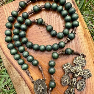 Puede incluir: Un rosario de madera verde con un crucifijo de bronce y una medalla de la Virgen María. Las cuentas del rosario son de color verde oscuro y están ensartadas en un cordón marrón. El crucifijo tiene la inscripción "JESUS NAZARENUS REX JUDÆORUM".
