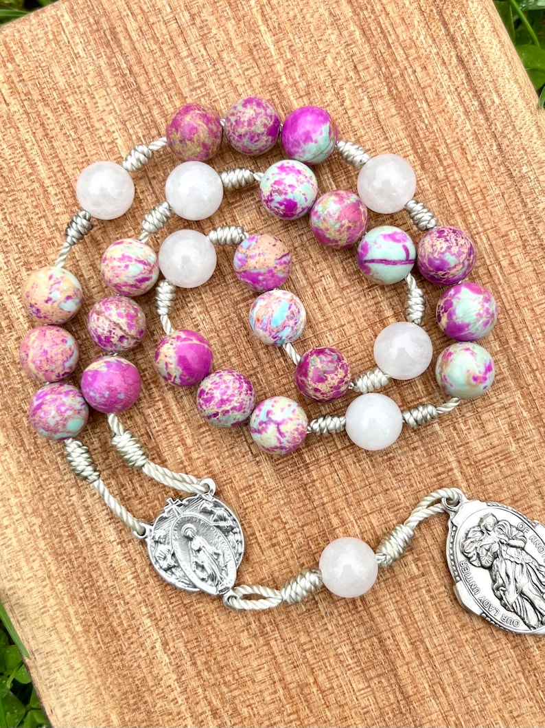 May include: A silver rosary with pink and white beads. The beads are made of a marbled stone. The rosary has a silver pendant with a depiction of the Virgin Mary and a silver pendant with a depiction of an angel.