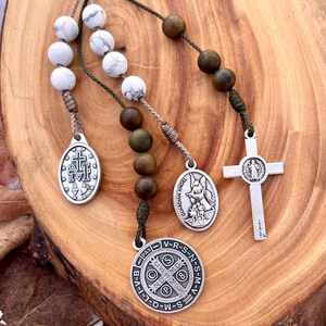 May include: A green and white rosary with silver charms. The charms include a cross, a guardian angel, and a medal with the words "V-R-S-N-S-M-V-I-I-D-W-S-S-N-M-V-R-S-N-S-M-V-I-I-D-W-S-S" around the edge.