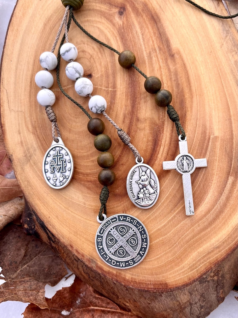 May include: A green and white rosary with silver charms. The charms include a cross, a guardian angel, and a medal with the words "V-R-S-N-S-M-V-I-I-D-W-S-S-N-M-V-R-S-N-S-M-V-I-I-D-W-S-S" around the edge.