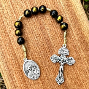 Könnte beinhalten: Ein Rosenkranz mit schwarzen und goldenen Perlen, einem silbernen Kruzifix und einem silbernen ovalen Anhänger. Das Kruzifix zeigt eine detaillierte Darstellung von Jesus. Der Anhänger hat ein Bild einer religiösen Figur. Die Perlen sind auf einer goldfarbenen Schnur aufgefädelt.
