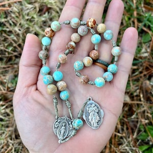 May include: A green and brown stone rosary with a silver charm featuring a religious image. The rosary is strung on a green cord and has a silver clasp.
