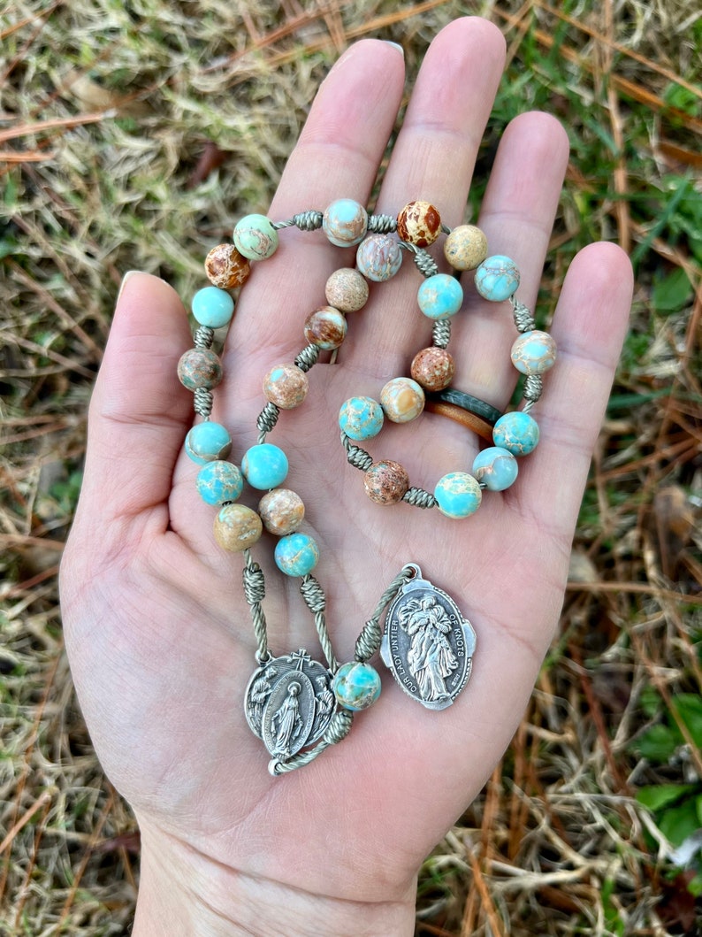 May include: A green and brown stone rosary with a silver charm featuring a religious image. The rosary is strung on a green cord and has a silver clasp.