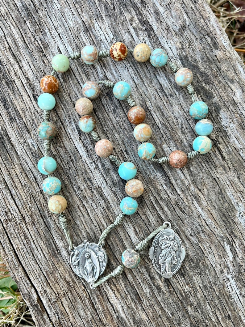 May include: A turquoise and brown stone rosary with silver beads and a silver pendant featuring the image of Our Lady of the Angels.