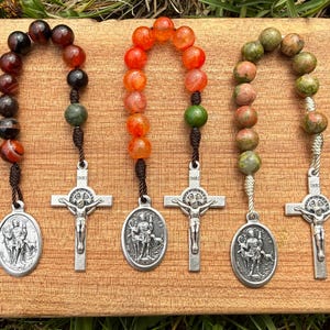 May include: Three rosaries with different colored beads: red, orange, and green. Each rosary has a silver crucifix and a silver pendant. The rosaries are displayed on a wooden surface.